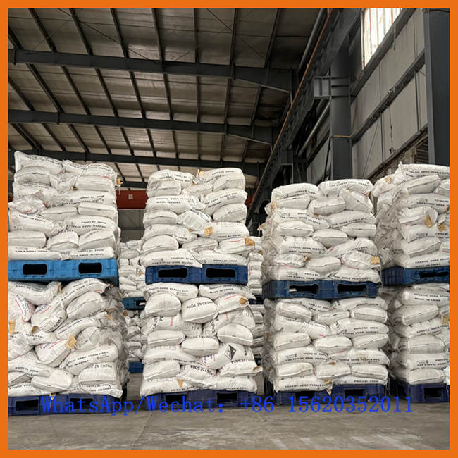 De grandes piles de sacs de perles de soude caustique de 25 kg stockés sur des palettes bleues dans un entrepôt. Les sacs portent les mentions 'MADE IN CHINA' et 'CAUSTIC SODA PEARLS'. Des informations de contact Whatsapp/Wechat sont superposées.